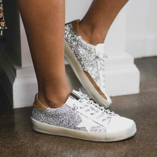 Trendy White Womens Sneakers with Silver Sparkly Sides Detailed with a White Star carved from the sparkles, a black thick line on the tongue of the shoe, and brown leather on the top of the very back of the sneaker.