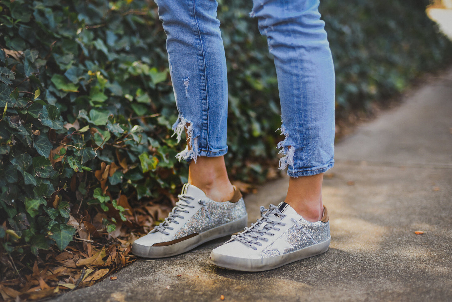Trendy White Womens Sneakers with Silver Sparkly Sides Detailed with a White Star carved from the sparkles, a black thick line on the tongue of the shoe, and brown leather on the top of the very back of the sneaker.